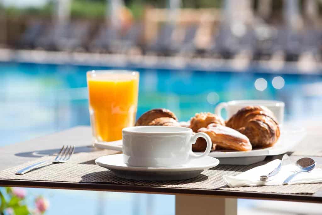 Café da manhã do hotel Insotel Club Maryland, Espanha, com uma xícara de café, um copo de suco de laranja e doces em uma mesa ao lado de uma piscina.