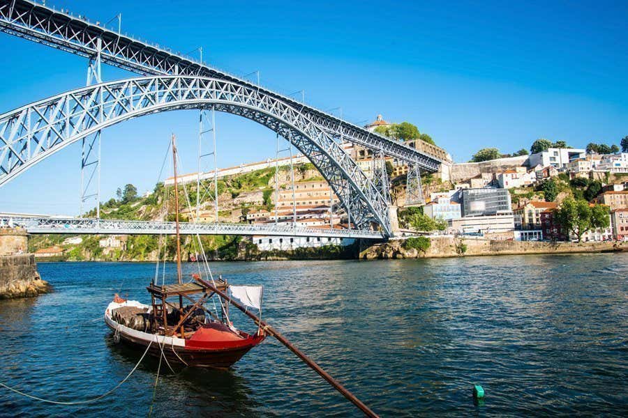 Um veleiro flutua em um rio sob uma grande ponte em arco de ferro, com edifícios e vegetação em uma colina ao fundo, representando o passeio Cruzeiro as sete pontes no Porto, em Portugal.