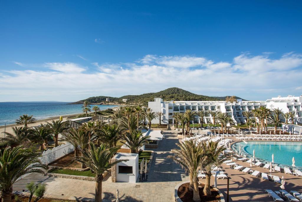 Fachada do Grand Palladium, Espanha, é um edifício branco à beira-mar com palmeiras, piscina externa, espreguiçadeiras e colinas ao fundo sob um céu azul, para representar hotéis em Ibiza.