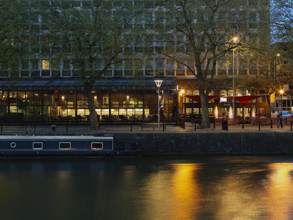 Fachada de vidro do The Bristol Hotel e assentos ao ar livre é visto em um canal calmo ao anoitecer, com luzes refletindo na água, para representar hotéis em Bristol.