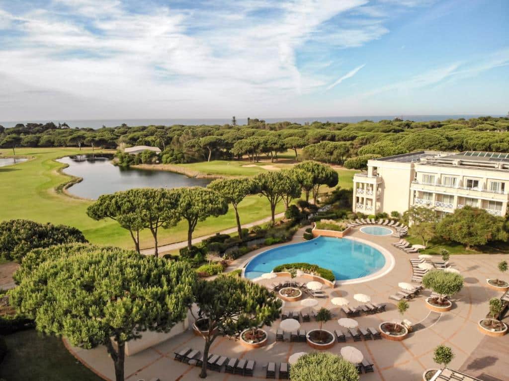 Hotel Onyria Quinta da Marinha, em Cascais com piscina é cercado por árvores, um campo de golfe e um lago sob um céu parcialmente nublado.