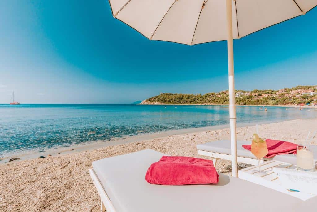Duas espreguiçadeiras com toalhas vermelhas sob um guarda-sol branco em uma praia de areia com mar azul e calmo, com bebidas em uma mesa, no Hotel La Bitta - Bovis Hotels. Representa hotéis em Sardenha.
